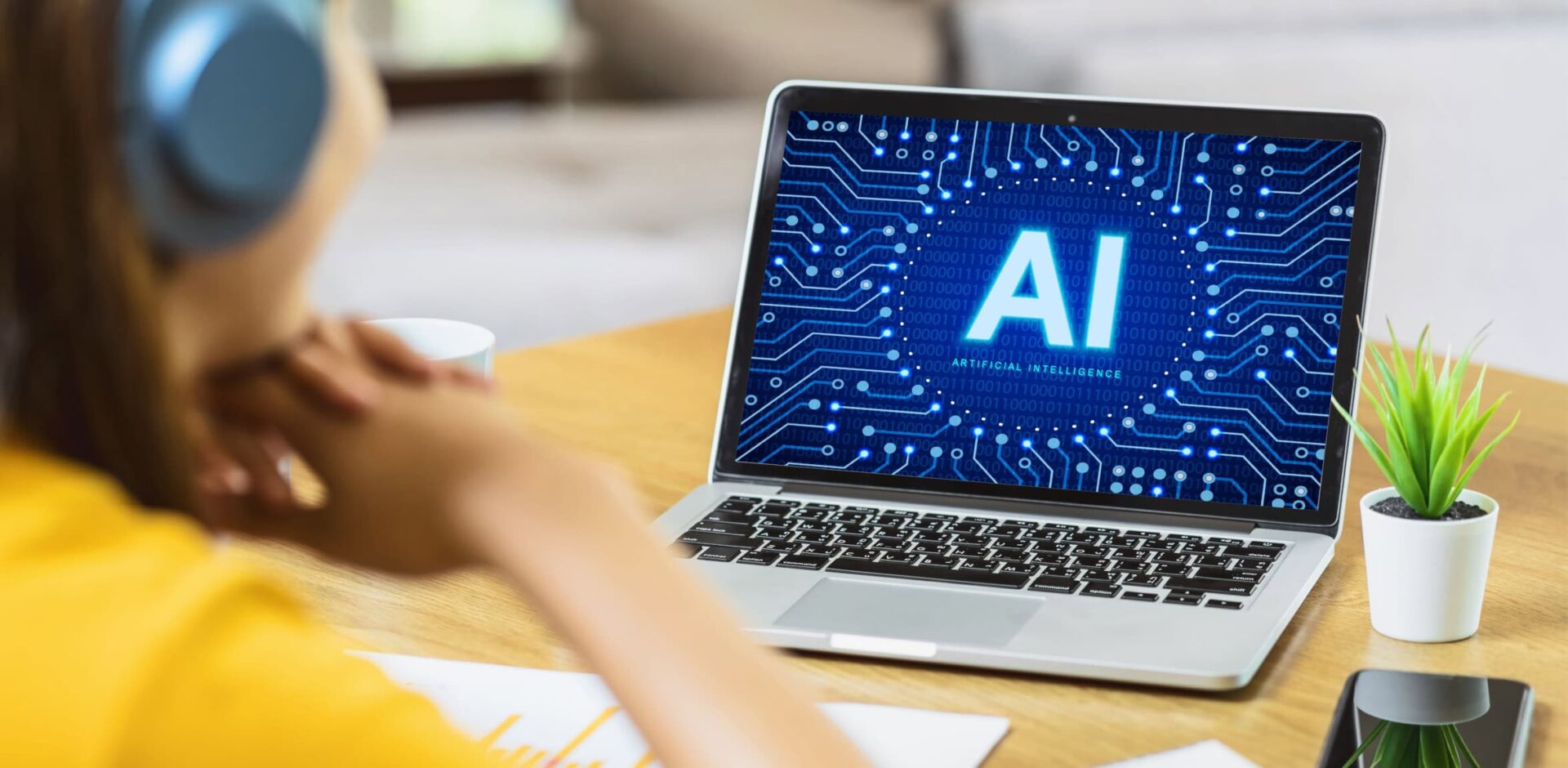 A young man is working and studying at his desk, using his laptop to log into an artificial intelligence website. The neon letters “AI” are displayed on his laptop screen. His plant and cell phone. By Jorge - AdobeStock